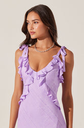 A woman is wearing a sleeveless, light purple dress with shoulder straps tied into bows. The dress features ruffles along the neckline and straps, and has a subtle textured pattern. She accessorizes with a silver chain necklace.